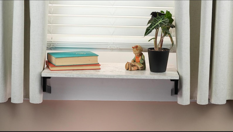 May include: A white shelf with black brackets is mounted above a window. The shelf is decorated with a small figurine of a girl reading a book, a potted plant, and three books.