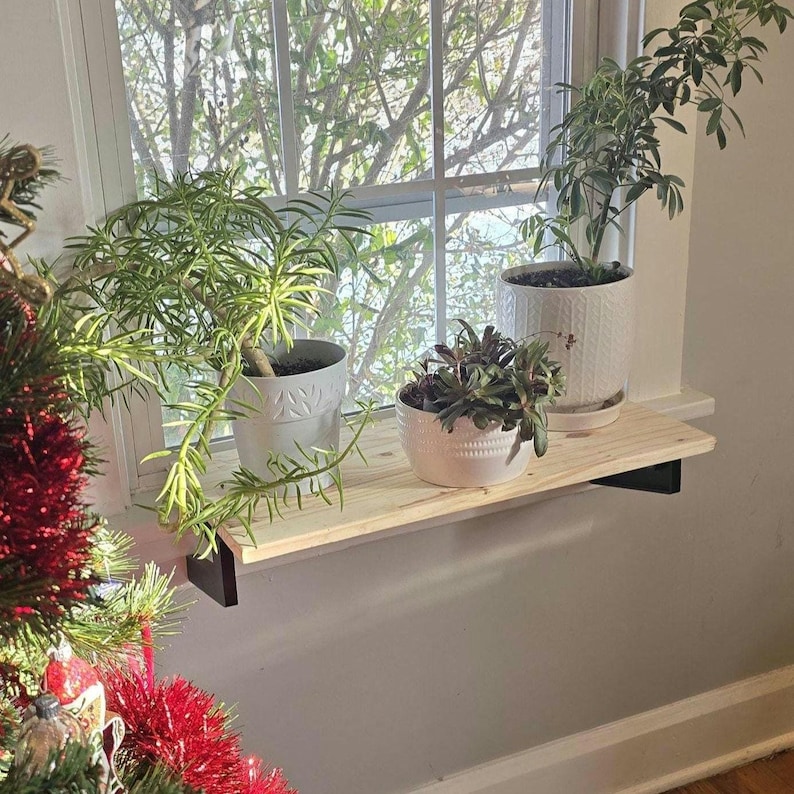 May include: A wooden shelf with three potted plants. The shelf is mounted on a white wall next to a window. The plants are in white ceramic pots. The shelf is made of natural wood and has a rustic look.