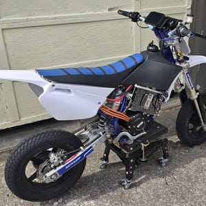 May include: A custom-built electric motorcycle with a white and blue frame, black tires, and a blue seat. The motorcycle is on a black stand with wheels. The tires have a Dunlop logo.