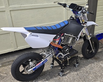 May include: A custom-built electric motorcycle with a white and blue frame, black tires, and a blue seat. The motorcycle is on a black stand with wheels. The tires have a Dunlop logo.