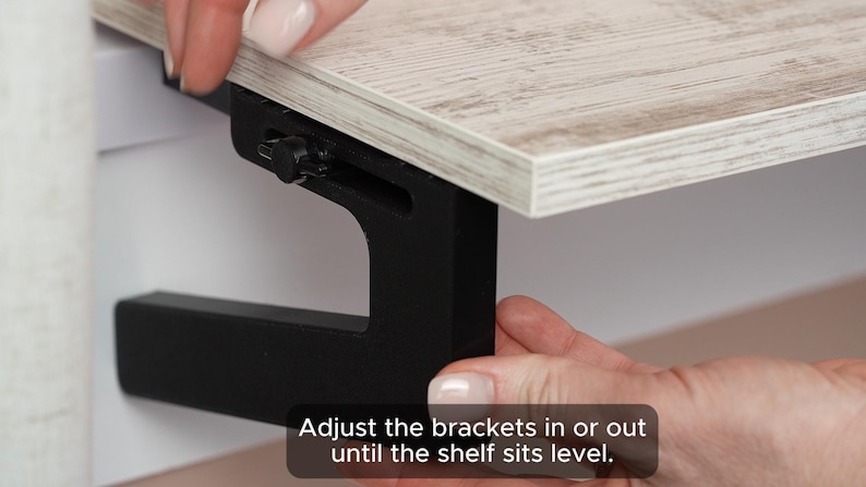 May include: A close-up of a black metal shelf bracket being adjusted to level a wooden shelf. The text "Adjust the brackets in or out until the shelf sits level." is visible in the image.