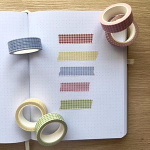May include: An open notebook with dotted pages, featuring several rolls of decorative washi tape in various colors and patterns. The tape is in shades of blue, red, yellow, pink, and green, with a gingham check design. Some tape is applied to the page.
