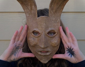 May include: A person wearing a brown paper mache mask with rabbit ears. The mask has a human-like face with large eyes and a neutral expression. The person is wearing a black shirt and has tattoos on their hands.