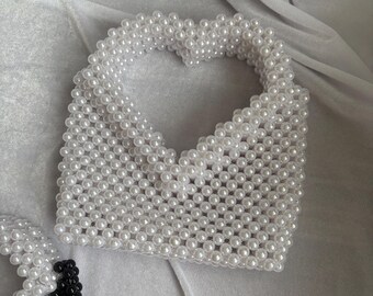 Puede incluir: Un bolso hecho de cuentas esféricas blancas dispuestas en un patrón de cuadrícula, con un recorte en forma de corazón en la parte superior que sirve como asa integrada. La estructura es uniforme, con cada cuenta unida para formar una superficie.