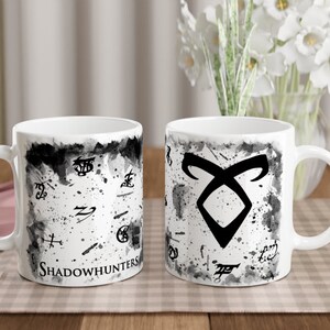 Könnte beinhalten: Zwei weiße Keramikbecher mit schwarz-weißen Designs. Ein Becher zeigt das Shadowhunters-Symbol und den Text "Shadowhunters". Der andere Becher zeigt eine Vielzahl von Runen.