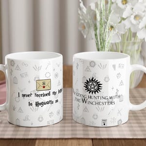 May include: Two white ceramic mugs with black text and illustrations. One mug reads "I never received my letter to Hogwarts so..." and the other says "I'm going hunting with the Winchesters". Both mugs have a handle and are on a checkered surface.