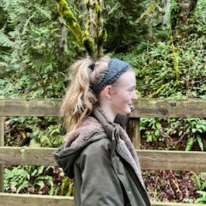 May include: A young person with long blonde hair wearing a gray headband, a brown hooded jacket, and a brown scarf standing on a wooden bridge in a forest.