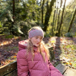 Peut inclure: Une jeune personne portant une veste matelassée rose avec une capuche doublée de fourrure est assise sur un banc en bois. Elle porte un bandeau tricoté rose et gris. L'arrière-plan est une image floue d'arbres.