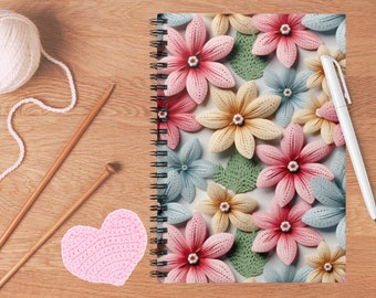 Pode incluir: Um caderno de espiral com um padrão floral de flores de papel quilling rosas, amarelas e azul-claras com folhas verdes. Ao lado do caderno estão duas agulhas de tricô de madeira, um novelo de lã branca, um coração de crochê rosa e uma caneta branca.