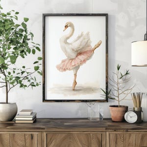 May include: A framed watercolour print of a swan in a ballet pose, wearing a pink tutu. The artwork is displayed on a wooden cabinet, with potted plants and decorative items. The swan is depicted with delicate brushstrokes.