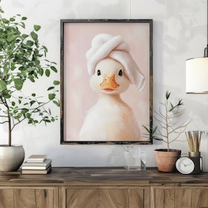 May include: Framed artwork featuring a charming duckling wearing a white towel turban. The duckling has a yellow beak and dark eyes, set against a soft pink background. The artwork is displayed on a wooden cabinet.