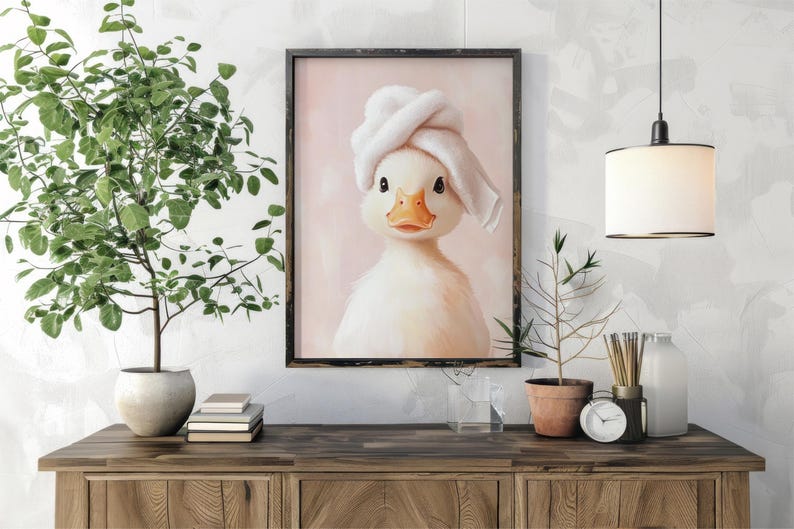 May include: Framed artwork featuring a charming duckling wearing a white towel turban. The duckling has a yellow beak and dark eyes, set against a soft pink background. The artwork is displayed on a wooden cabinet.