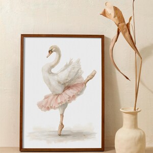 May include: Watercolour painting of a graceful white swan in a pink tutu, en pointe. The artwork is framed in a dark wood frame. The swan is depicted in a ballet pose. The artwork is displayed on a wooden surface.