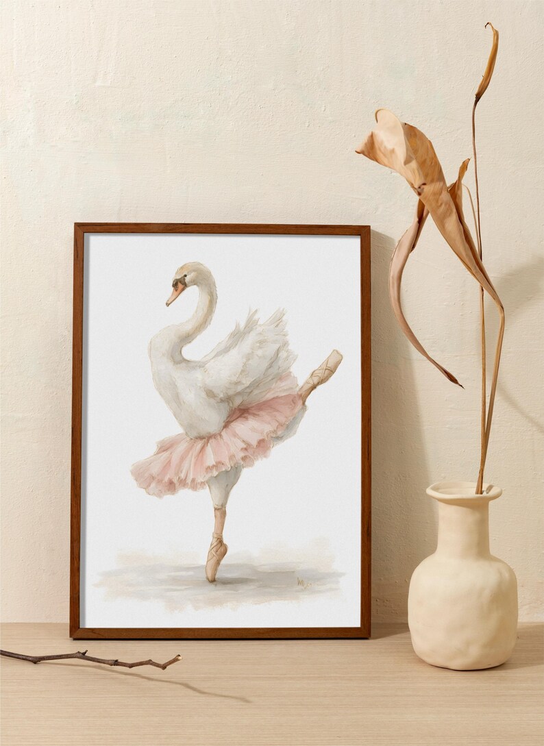 May include: Watercolour painting of a graceful white swan in a pink tutu, en pointe. The artwork is framed in a dark wood frame. The swan is depicted in a ballet pose. The artwork is displayed on a wooden surface.