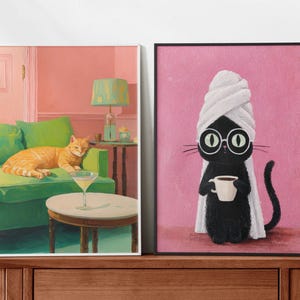 May include: Two framed art prints. One shows an orange cat on a green sofa with a martini. The other depicts a black cat wearing glasses and a towel, holding a coffee cup. The backgrounds are pink.