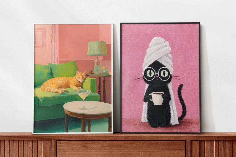 May include: Two framed art prints. One shows an orange cat on a green sofa with a martini. The other depicts a black cat wearing glasses and a towel, holding a coffee cup. The backgrounds are pink.