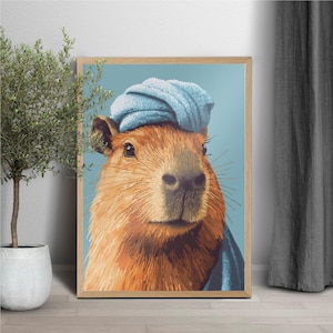 May include: A framed art print featuring a capybara wearing a blue towel on its head. The capybara has brown fur and is set against a light blue background. The artwork is in a wooden frame.