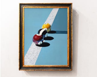 May include: Framed painting of three billiard balls on a light blue background with a white stripe. The balls are red, black and white, and yellow. The frame is black with gold trim.