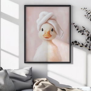 May include: A framed print featuring a charming duckling wearing a white towel on its head. The duckling has a yellow beak and is set against a soft pink background. The artwork is displayed in a black frame.