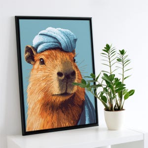May include: A framed art print featuring a capybara wearing a blue towel on its head. The capybara has brown fur and a detailed face. The background is a light blue, and a small potted plant sits to the right of the frame. The frame is black.