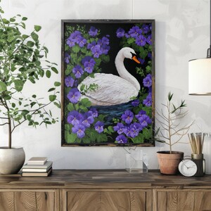 Op de afbeelding: Een ingelijst schilderij van een witte zwaan met een oranje snavel, omringd door paarse bloemen en groene bladeren. Het kunstwerk wordt tentoongesteld op een houten kast, met een plant en andere decoratieve items.