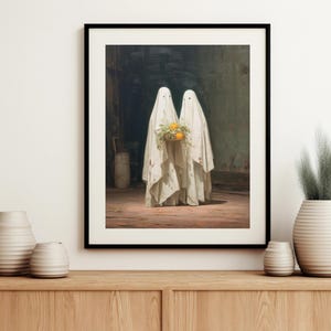 May include: Framed art print featuring two figures draped in white sheets, resembling ghosts, holding a bouquet of orange flowers. The artwork has a vintage aesthetic, set against a dark, moody background.