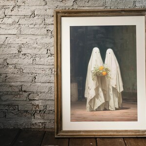 May include: Framed artwork featuring two figures draped in white sheets, resembling ghosts. They hold a bouquet of yellow-orange flowers. The artwork is in a gold-toned frame, set against a brick wall and wooden floor.