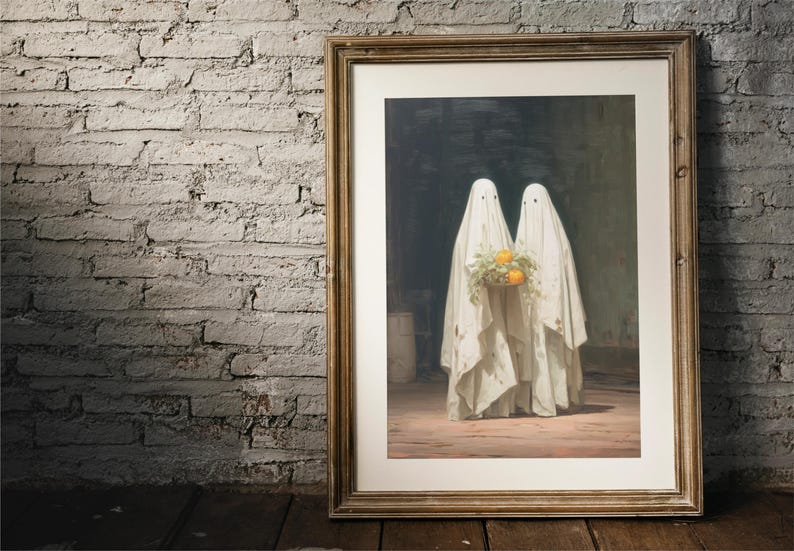 May include: Framed artwork featuring two figures draped in white sheets, resembling ghosts. They hold a bouquet of yellow-orange flowers. The artwork is in a gold-toned frame, set against a brick wall and wooden floor.