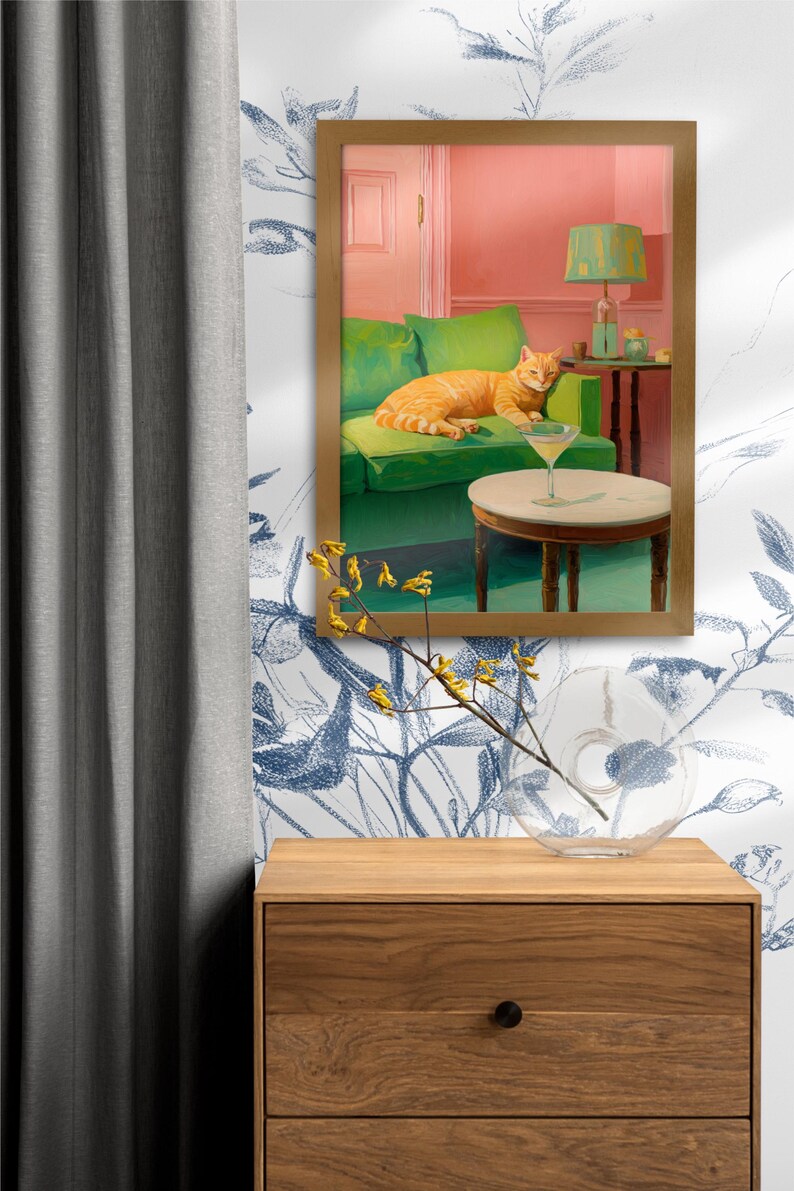 May include: A framed painting shows an orange tabby cat lounging on a green sofa, with a martini on a side table. The room has a pink wall and a green lamp. A wooden chest of drawers and a glass vase are in the foreground.