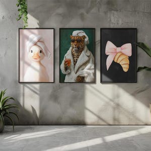 May include: Three framed art prints hang on a gray wall. The prints feature a duck wearing a towel, a leopard in a robe, and a croissant with a pink bow. A potted plant sits on the floor to the right, and a window is on the left.