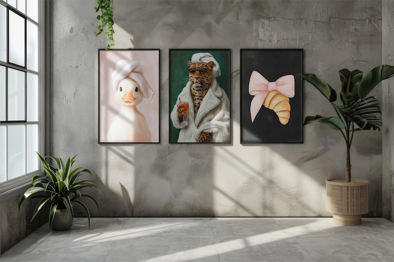 May include: Three framed art prints hang on a gray wall. The prints feature a duck wearing a towel, a leopard in a robe, and a croissant with a pink bow. A potted plant sits on the floor to the right, and a window is on the left.