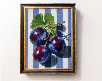 May include: A framed still life painting of three ripe plums with a green leaf and stem. The plums are a deep blue with red highlights, set against a blue and white striped background. The frame is black with a gold trim.