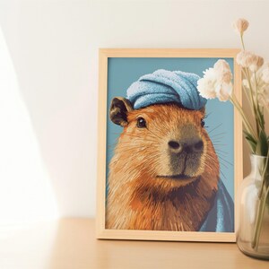 May include: A framed art print featuring a capybara wearing a blue towel on its head. The capybara has brown fur and is set against a light blue background. A vase of dried flowers sits to the right.