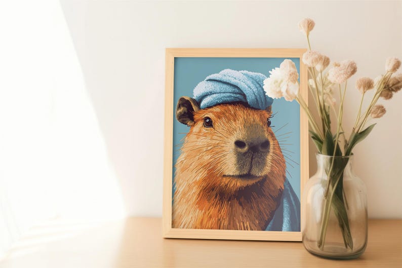 May include: A framed art print featuring a capybara wearing a blue towel on its head. The capybara has brown fur and is set against a light blue background. A vase of dried flowers sits to the right.