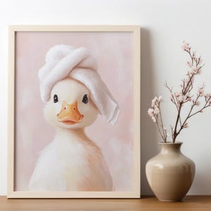 May include: A framed print featuring a charming duckling wearing a white towel on its head. The duckling has a yellow beak and is set against a soft pink background. A vase with flowers is next to the frame.