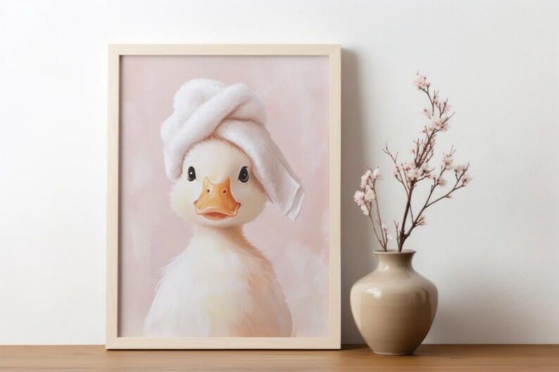 May include: A framed print featuring a charming duckling wearing a white towel on its head. The duckling has a yellow beak and is set against a soft pink background. A vase with flowers is next to the frame.