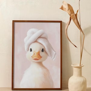 May include: A framed print featuring a charming white duckling wearing a white towel on its head. The duckling has a yellow beak and is set against a soft pink background. The frame is a dark brown color. A vase with dried plants is on the right.
