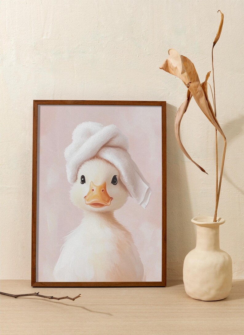 May include: A framed print featuring a charming white duckling wearing a white towel on its head. The duckling has a yellow beak and is set against a soft pink background. The frame is a dark brown color. A vase with dried plants is on the right.