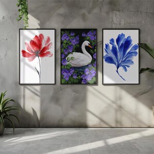 Op de afbeelding: Drie ingelijste aquarelprints. De prints tonen een rode bloem, een witte zwaan omringd door paarse bloemen en een blauw abstract bloemmotief. Het kunstwerk wordt tentoongesteld op een grijze muur met een raam en planten.