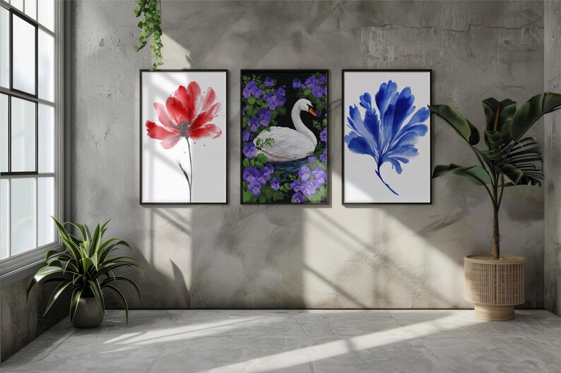 Op de afbeelding: Drie ingelijste aquarelprints. De prints tonen een rode bloem, een witte zwaan omringd door paarse bloemen en een blauw abstract bloemmotief. Het kunstwerk wordt tentoongesteld op een grijze muur met een raam en planten.