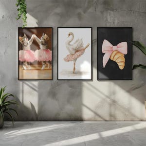 May include: Three framed art prints with ballet themes. One print shows two cats in pink tutus, another a swan in a tutu, and the third a croissant with a pink bow. The prints are displayed on a grey wall.