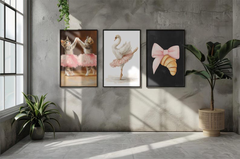 May include: Three framed art prints with ballet themes. One print shows two cats in pink tutus, another a swan in a tutu, and the third a croissant with a pink bow. The prints are displayed on a grey wall.