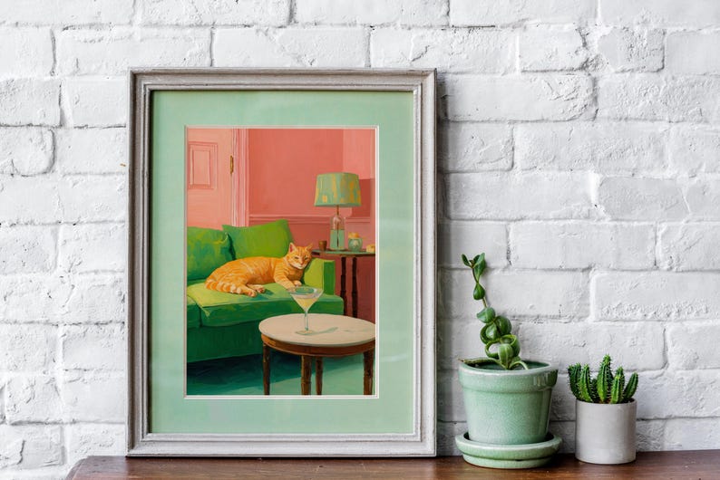 May include: Art print in a light green frame depicting an orange tabby cat on a green sofa. A cocktail sits on a small table. The room has a pink wall and a green lamp. The artwork is a stylish interior scene.