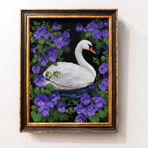 Op de afbeelding: Een ingelijst kunstwerk met een witte zwaan met een oranje snavel, omringd door paarse bloemen en groene bladeren tegen een zwarte achtergrond. Het kunstwerk zit in een sierlijke goud- en zwarte lijst.
