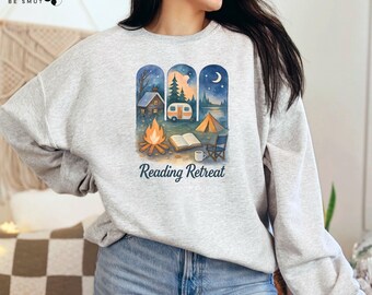 May include: A light gray sweatshirt with a watercolor design featuring a camping scene with a cabin, camper, tent, and campfire. The text "Reading Retreat" is below the image. The sweatshirt is made of a soft material.