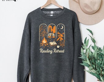 May include: A dark grey sweatshirt with a camping-themed graphic. The design includes a cabin, camper van, tent, campfire, and open book, in orange and white. The words "Reading Retreat" are printed below the graphic.