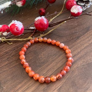 Peut inclure: Un bracelet extensible composé de perles de pierres précieuses orange et rouge. Le bracelet circulaire repose sur une surface en bois. L'arrière-plan comprend des baies rouges et des pommes de pin, suggérant un thème de vacances.