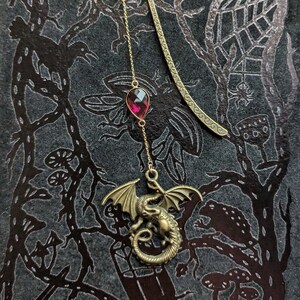May include: A gold bookmark with a dragon design and a dangling red gemstone. The bookmark is on a black background with a silver pattern.