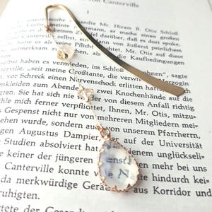 Könnte beinhalten: Ein goldenes Lesezeichen mit einem klaren Glasanhänger, auf dem in Kursivschrift "Das Gespenst von Canterville" steht. Das Lesezeichen ist mit kleinen weißen Perlen verziert.