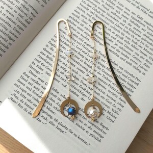 Puede incluir: Dos clips de marcapáginas de oro con abalorios decorativos. Un abalorio es una luna creciente con una piedra preciosa azul y el otro es una luna creciente con una perla. Los marcapáginas están en un libro con texto.
